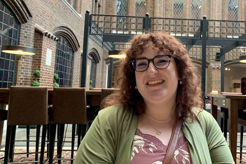 Christiana Haight Delta College student sits at a table in a courtyard smiling for a photo. She wears black-rimmed glasses a light green cardigan and has red-brown curly hair