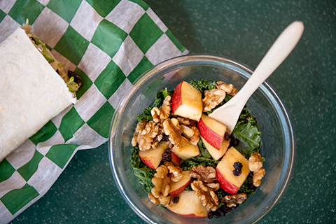 glass bowl of diced apples, raisins, kale and walnuts with a spoon and a sandwich wrap to the left of it