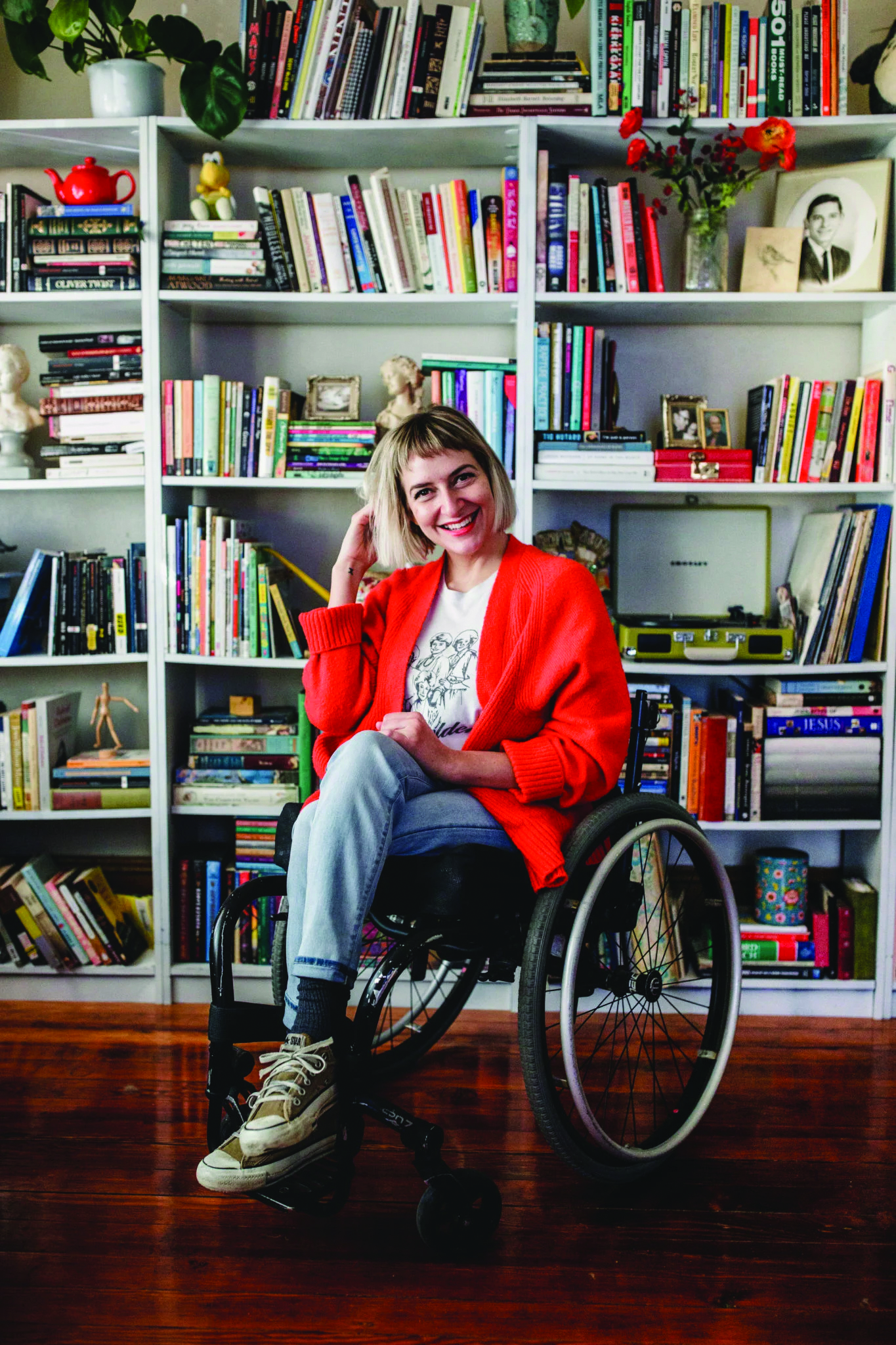 white female presenting woman with chin-lengthed blond hair sits smiling with her legs crossed in a manual wheelchair positioned infront of a bookcase